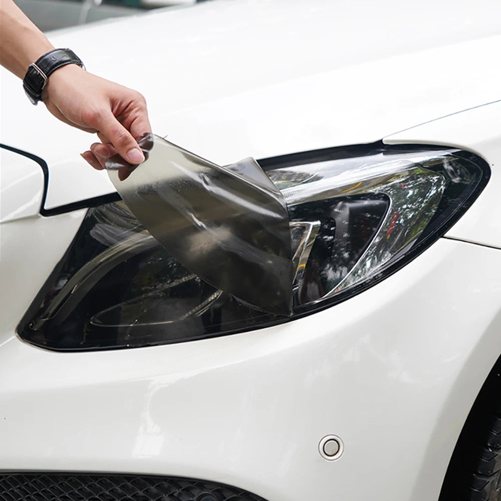 A hand is applying a smoke tint vinyl decal to the headlight of a white car.