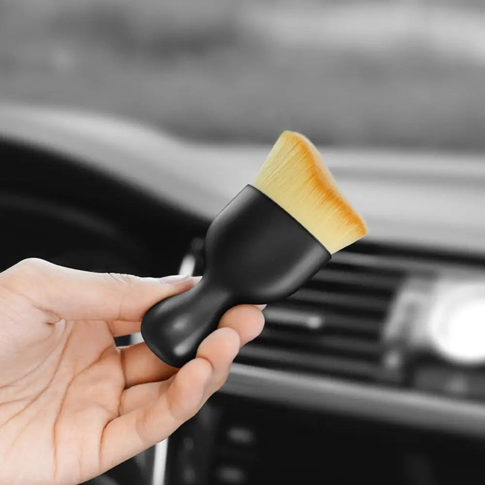 A hand holds a black silicone car interior cleaning brush with a yellow bristles, positioned over a car vent.
