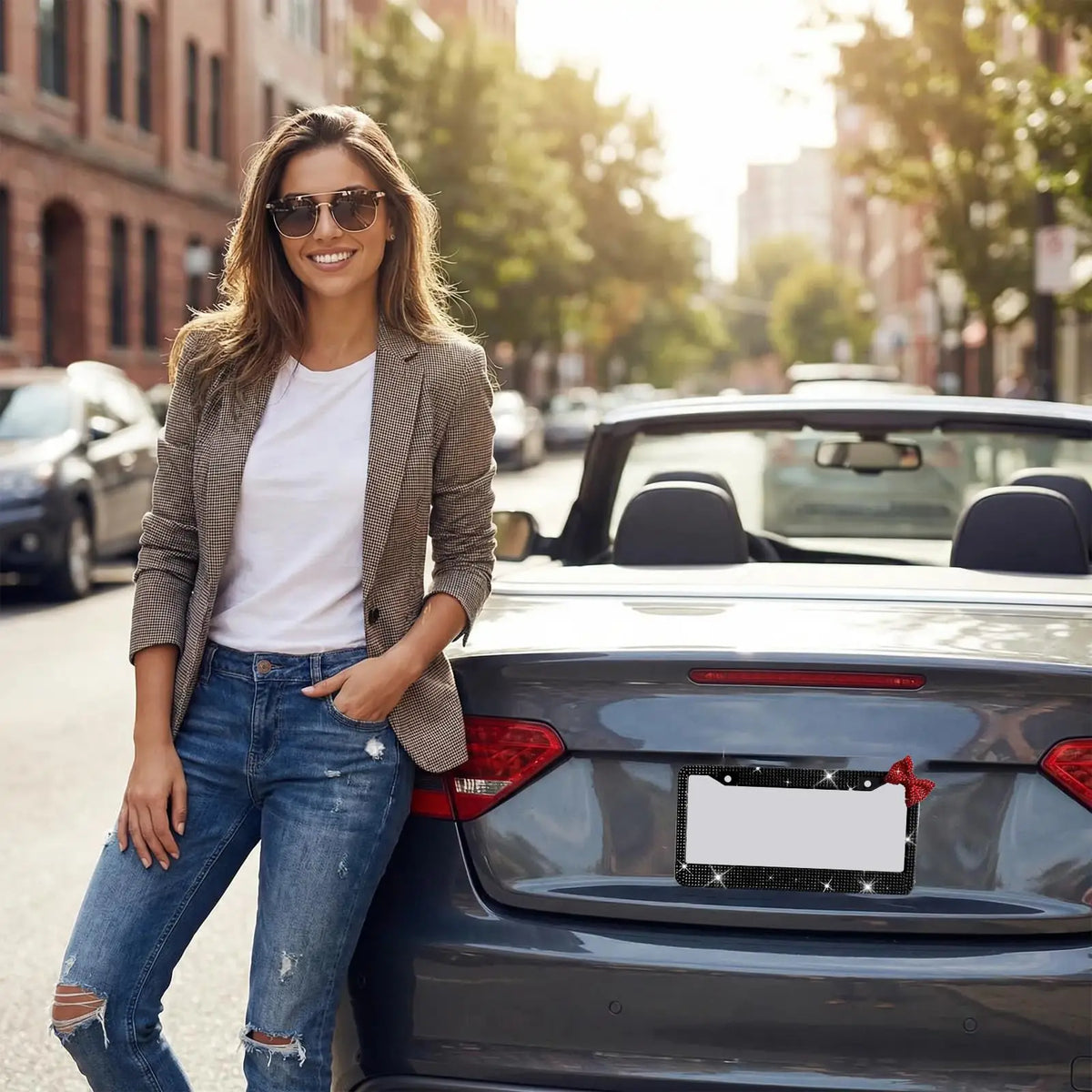 A young woman smiling while standing next to a dark gray car with a decorative license plate frame set featuring a red bow ac