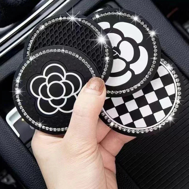 A hand holds a set of two black silicone car cup coasters with white floral designs and silver glitter edges.