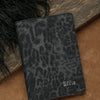 A black leopard print passport holder resting on a brown surface next to dark fur.