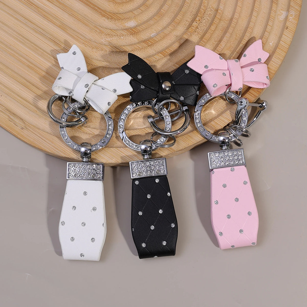 Drei Luxus-PU-Leder-Bow-Knoten-Schlüsselanhänger in Weiß, Schwarz und Pink auf Holzoberfläche.