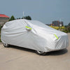 A white universal all-weather car cover is placed over a silver SUV or sedan in an outdoor parking lot.