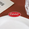 A red oval enamel lapel pin featuring the Peterbilt logo placed on a white ceramic plate.