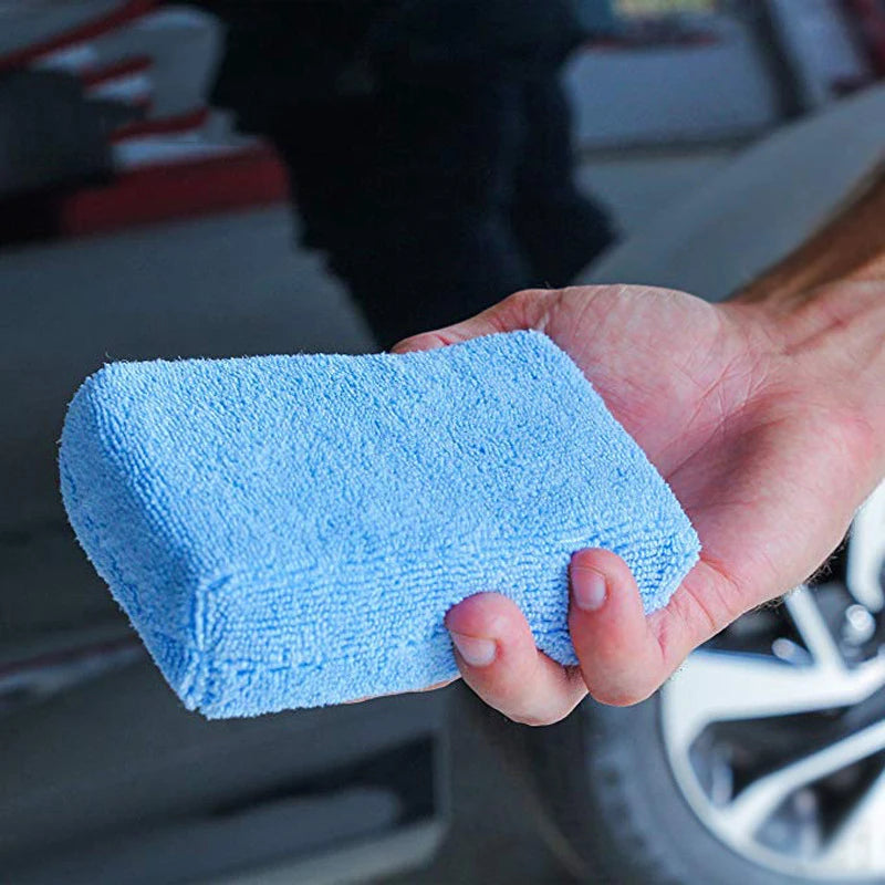 A hand holds a bright blue microfiber polishing sponge for car wax application.