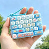 A hand holds a light blue keyboard with cartoon keycaps and a blue beaded keychain.