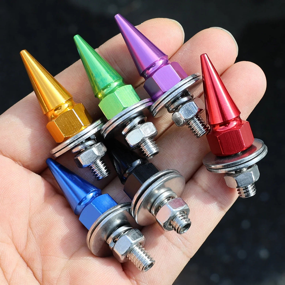 A hand holds several multi-colored M6 license plate fixing screws with conical heads and threaded bases.