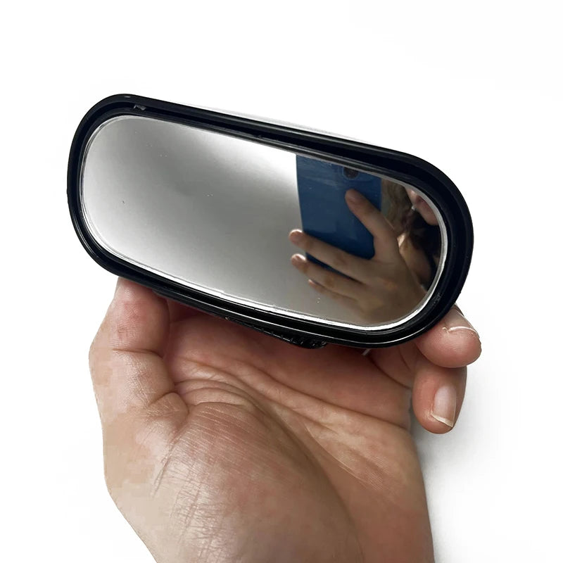 A hand holds a black, oval-shaped 360° adjustable car blind spot mirror reflecting the user's face.