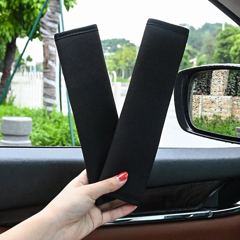A hand holds two black plush seat belt covers against a car window.