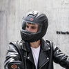 A man wearing a black motorcycle helmet and leather jacket is shown in a studio setting.