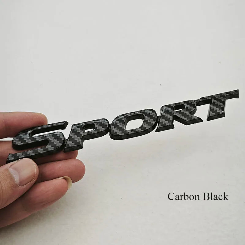 A hand holds a black carbon fiber logo emblem with the word SPORT engraved on it.