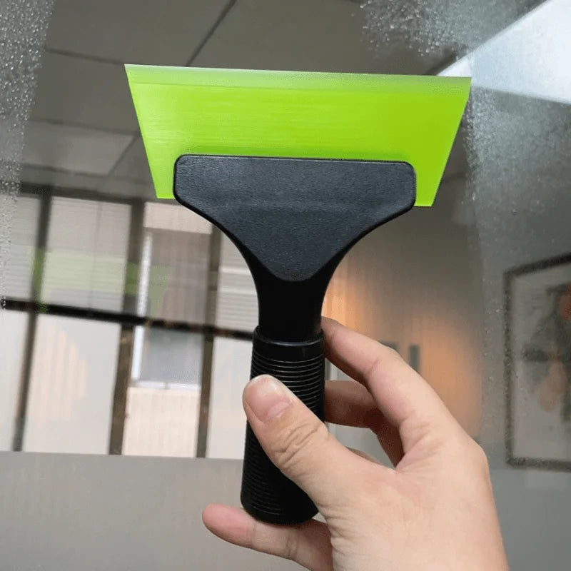 Eine Hand benutzt einen leuchtend limettengrünen unsichtbaren Autofilmschaber, um Wasser aus einem Fenster zu entfernen.