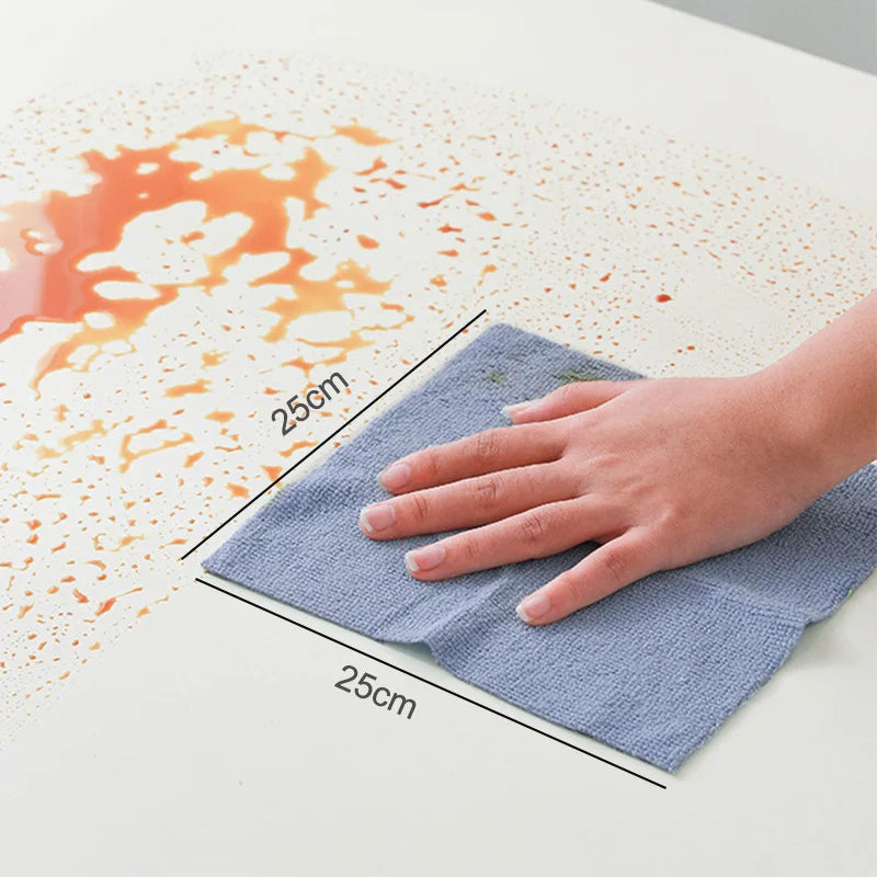 A hand is using a blue reusable microfiber cleaning towel to wipe up orange liquid stains on a white surface.