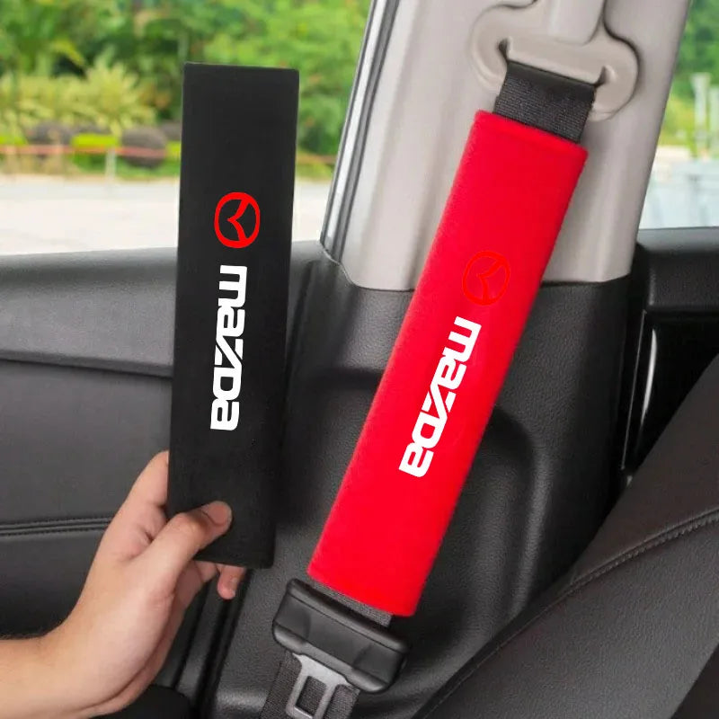 A red seat belt shoulder pad is shown attached to a car headrest, next to a black Mazda branded strap.