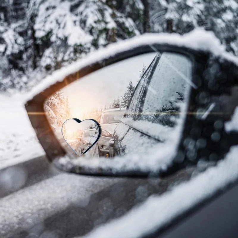 A heart-shaped car rearview mirror reflecting a snowy forest and bright light.