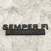 Black metal emblem with the text SEMPER FI EDITION on a light gray textured surface.