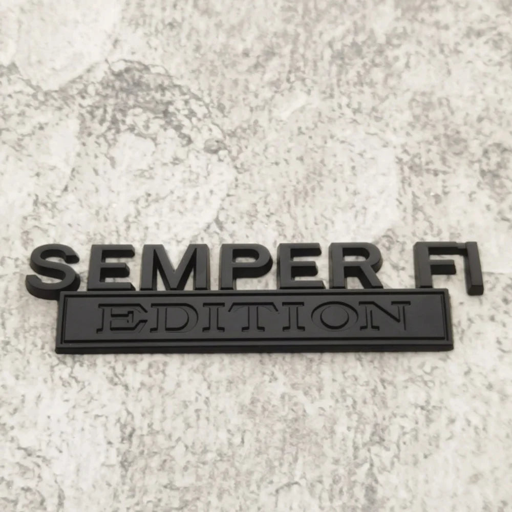 Black metal emblem with the text SEMPER FI EDITION on a light gray textured surface.