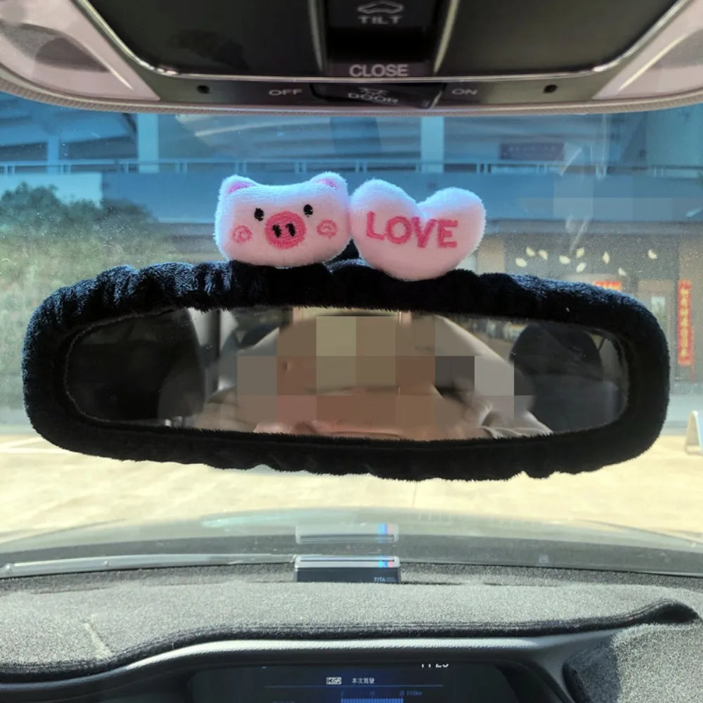 A pink pig and heart plush decoration placed on a black rearview mirror cover inside a car.