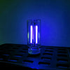 A clear glass cylindrical LED night light with a bright white glow sits on a black tray against a blue background.