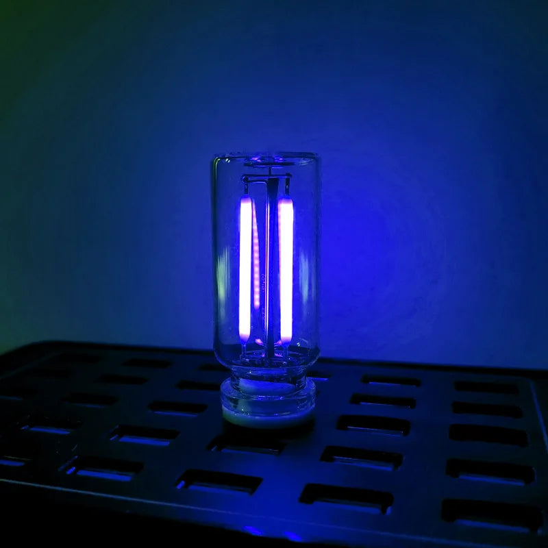 A clear glass cylindrical LED night light with a bright white glow sits on a black tray against a blue background.