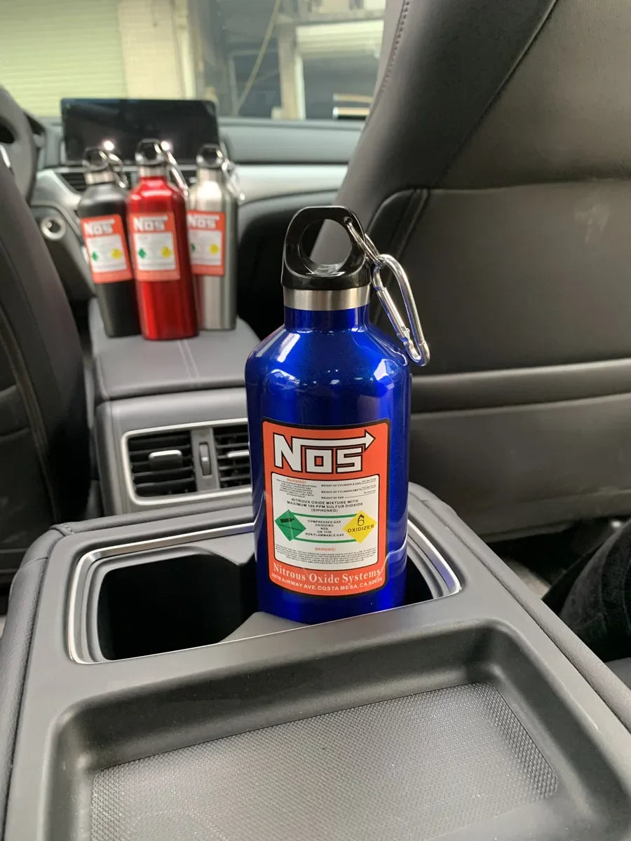 A blue insulated stainless steel travel kettle is placed in a car cup holder alongside several red and silver bottles.