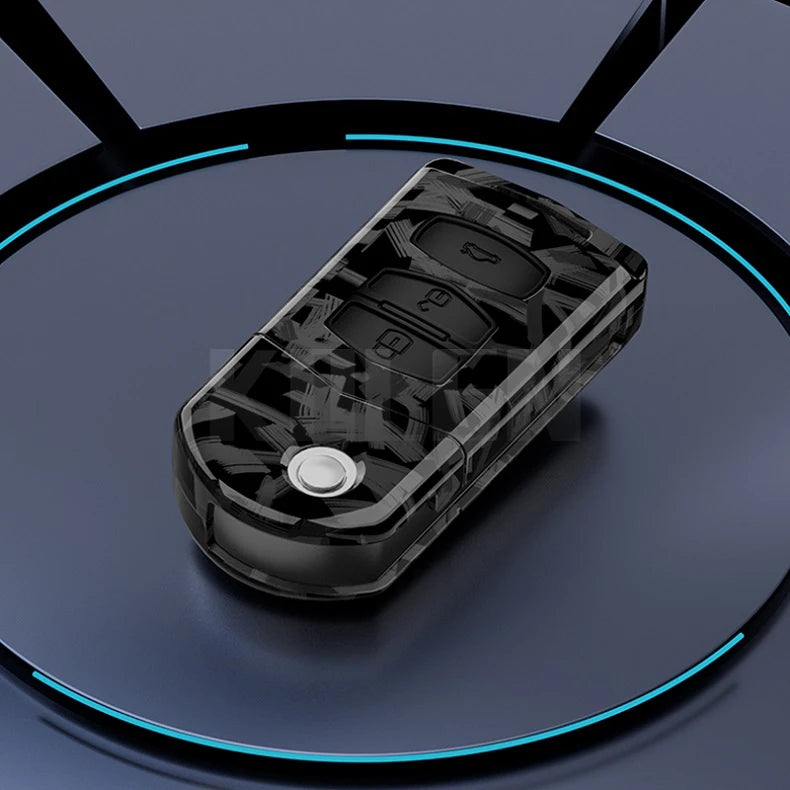 A black carbon fiber button cover for a car key is displayed on a dark surface.
