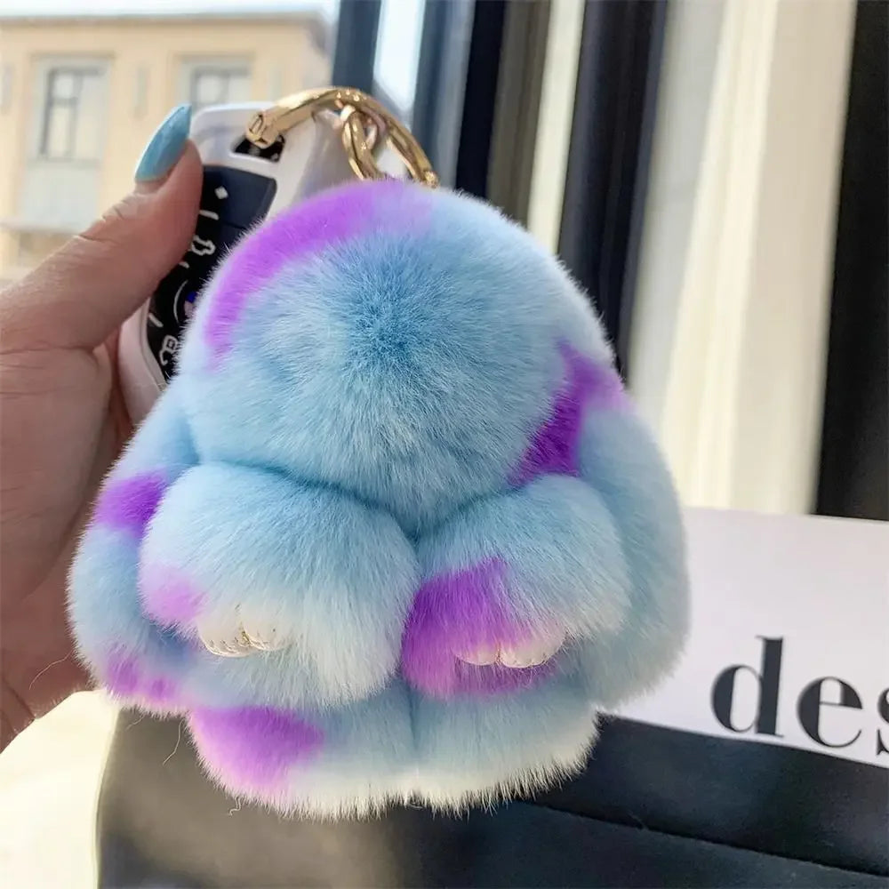 A light blue and purple plush bunny keychain with soft fur and a gold ring being held by a hand.