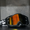 A pair of black high-performance motocross goggles with orange tinted lenses resting on a dark surface.
