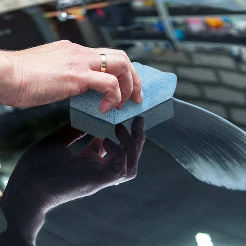 A hand uses a blue microfiber pad to polish a dark car surface.