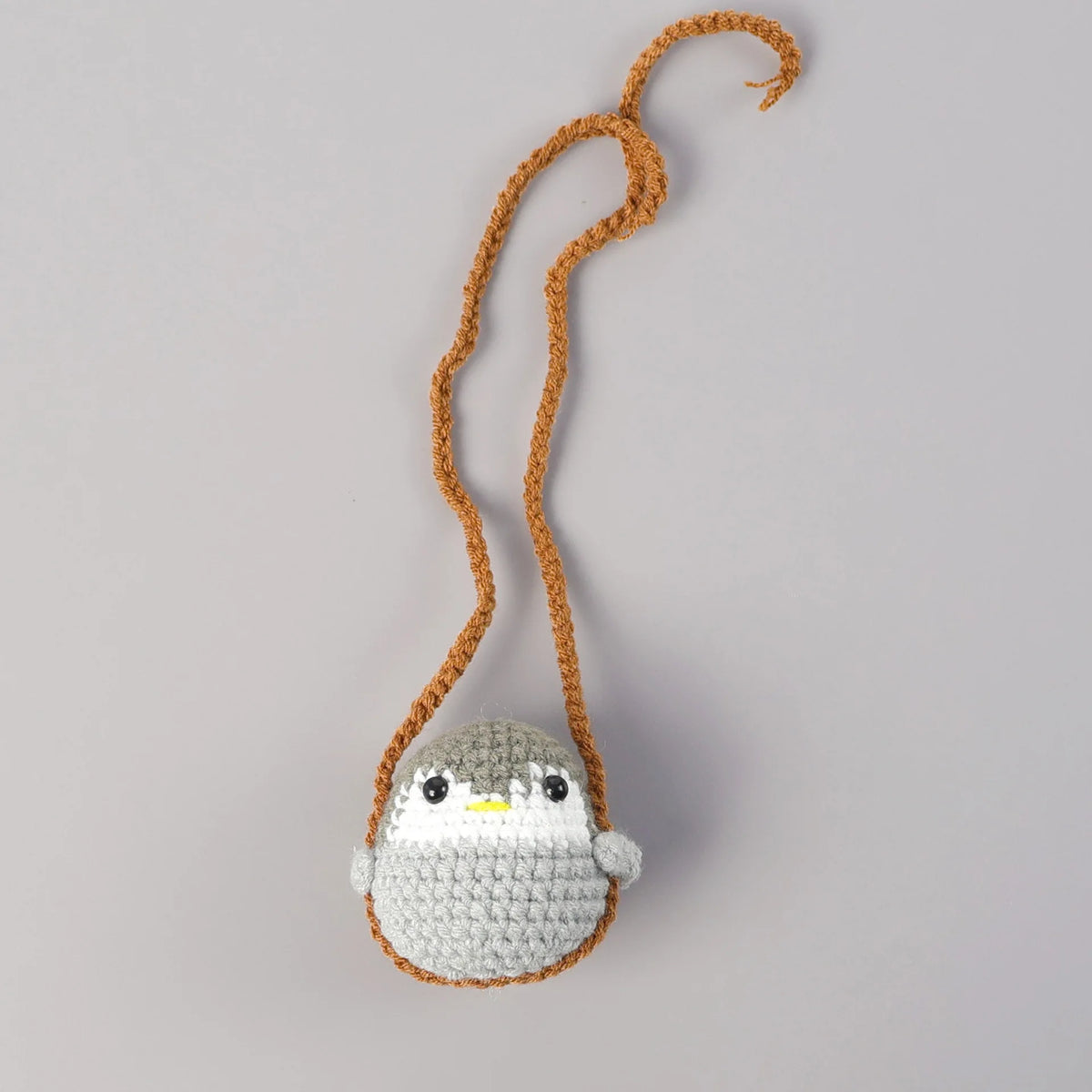 A crocheted hanging decor featuring a small, round bird with gray and white feathers, suspended by a brown string.