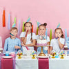 Ein Mädchen in einem weißen Hemd hält einen rosa Cupcake auf einem Ständer mit bunten Kegelpartyhüten.