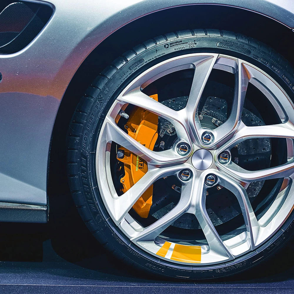 Close-up of a silver car wheel with an orange brake caliper and reflective safety stickers on the tire.
