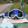 A pair of white and blue motocross goggles resting on a gray surface with a white cloth and a clear lens.