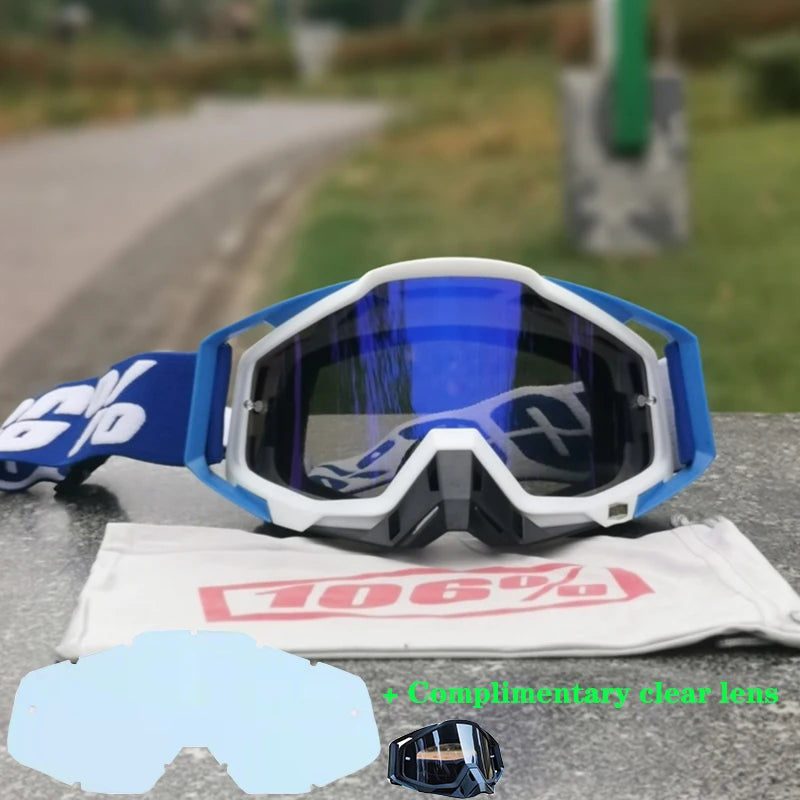 A pair of white and blue motocross goggles resting on a gray surface with a white cloth and a clear lens.