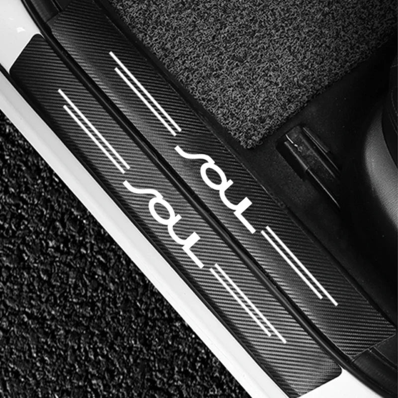 Close-up of a black carbon fiber door sill protector with white logo lines on a dark surface.