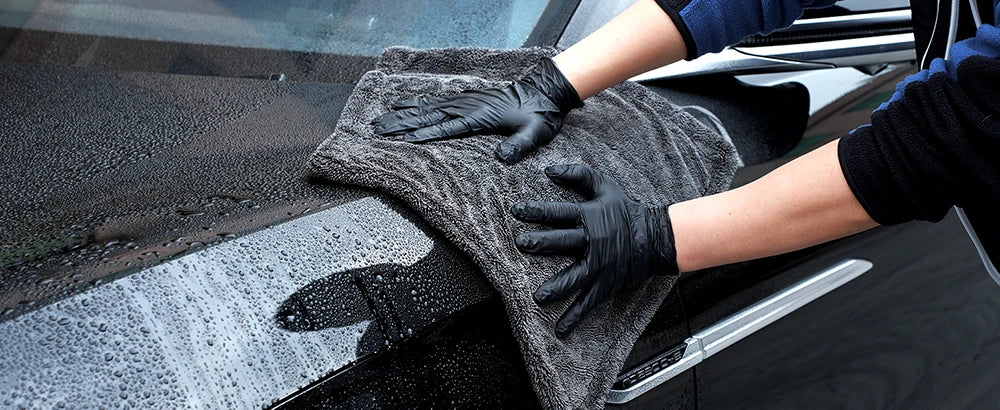 Hands wearing black gloves are cleaning a car window with a gray microfiber cloth.