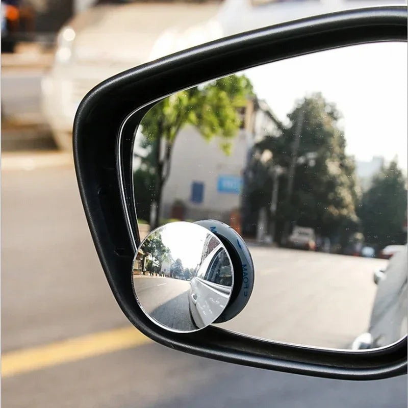 A close-up view of a black car rearview mirror with a circular blind spot camera mounted on it.