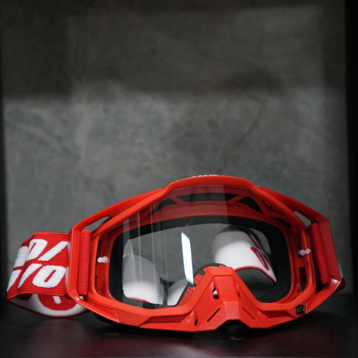 A pair of bright orange motocross goggles with a red frame and clear lenses resting on a dark surface.
