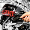 A hand is using a red and black wool cleaning brush to wash dirt from a silver wheel rim.