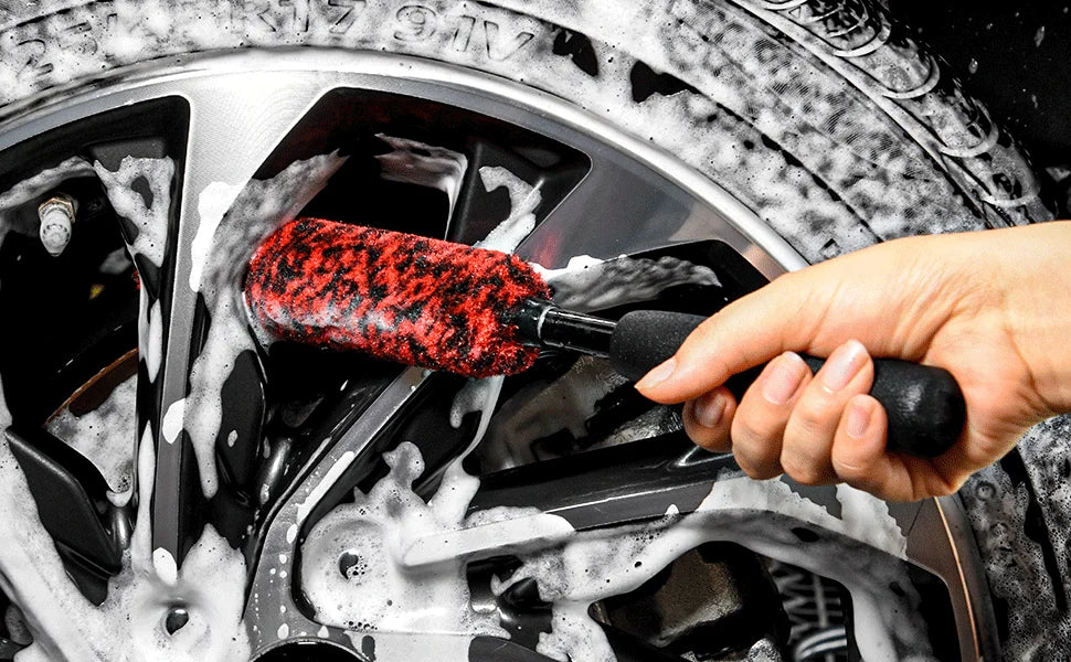A hand is using a red and black wool cleaning brush to wash dirt from a silver wheel rim.