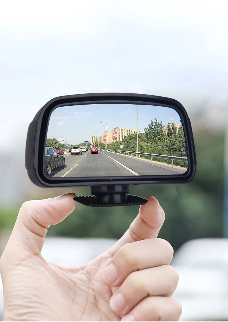 Une main tient un miroir convexe noir montrant une vue d'une route et de bâtiments environnants.