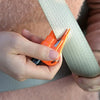 A hand holds an orange emergency car escape tool with a window breaker and seatbelt cutter against a beige strap.