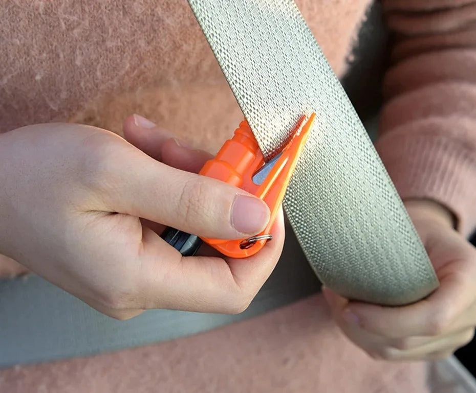 A hand holds an orange emergency car escape tool with a window breaker and seatbelt cutter against a beige strap.