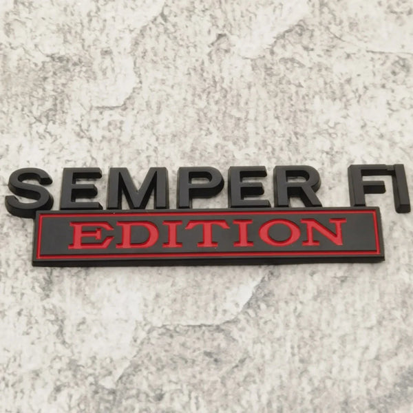 Black metal emblem with red lettering reading SEMPER FI EDITION on a gray marbled surface.