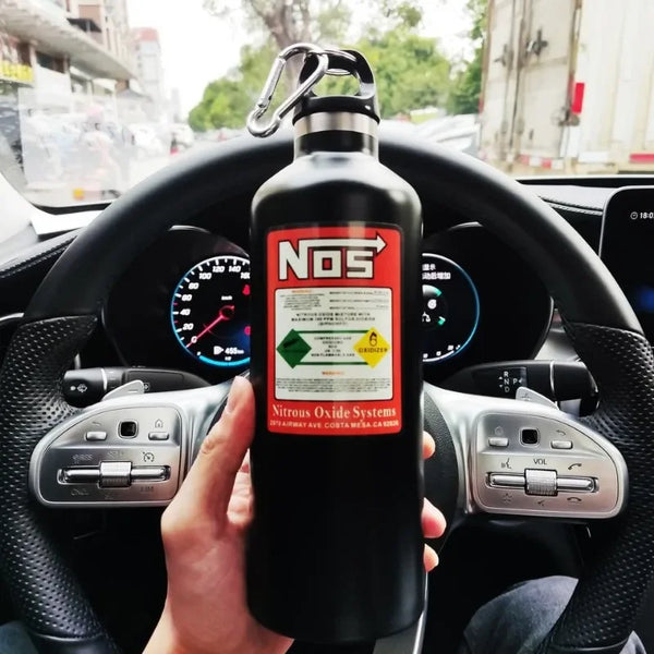 A hand holds a black insulated stainless steel travel kettle in the steering wheel of a car.