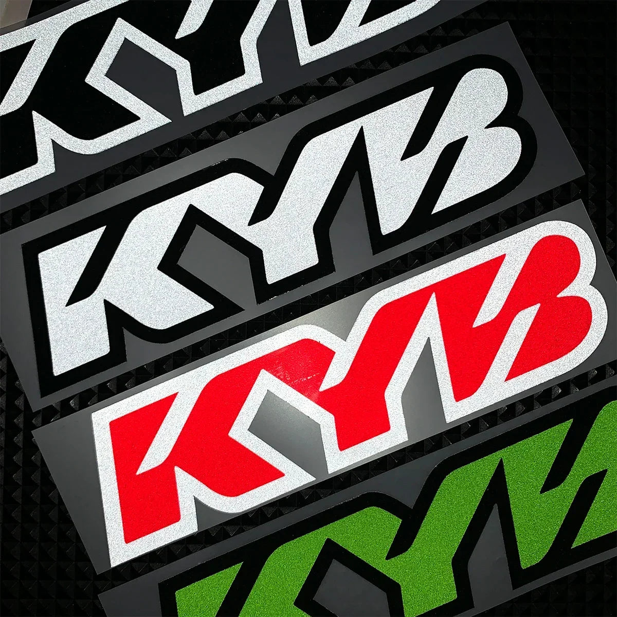 A close-up of black and white racing suspension decals featuring the KYB logo in various colors.