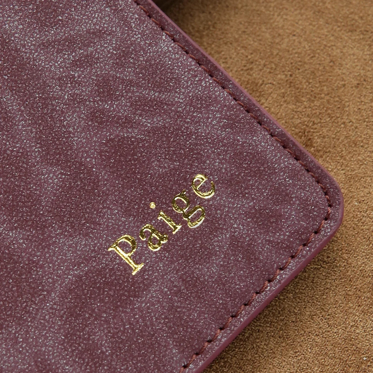 A close-up of a deep purple textured leather passport holder with gold embossed writing on a brown suede background.
