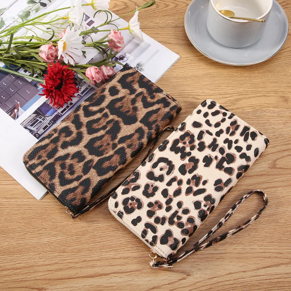 A leopard print long wallet resting on a wooden surface next to some flowers and a cup.