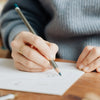 A person's hand writing on paper with a pencil, wearing a gray sweater.