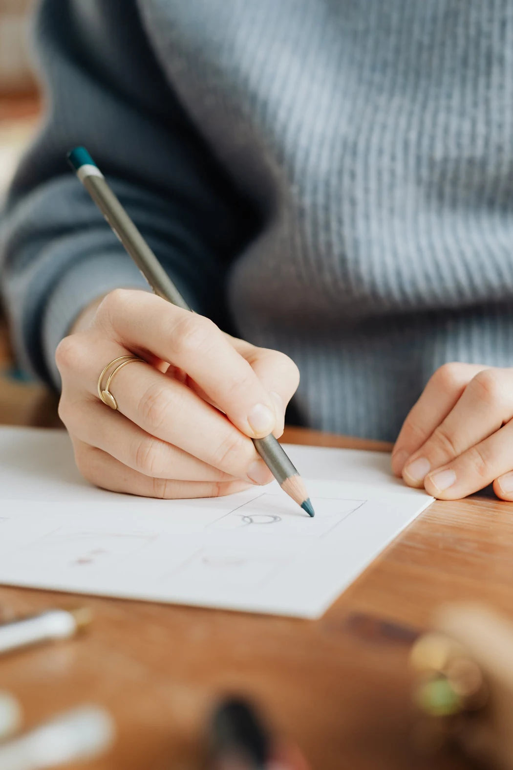 A person's hand writing on paper with a pencil, wearing a gray sweater.